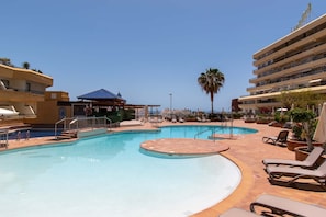 A swimming pool with sun loungers and a covered area, surrounded by buildings with balconies.