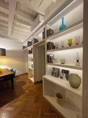 A modern interior with wooden herringbone flooring, white shelving units displaying books and decorative items, and a dining area with a wooden table and chairs. - Carpe Diem Sistina (Roma)