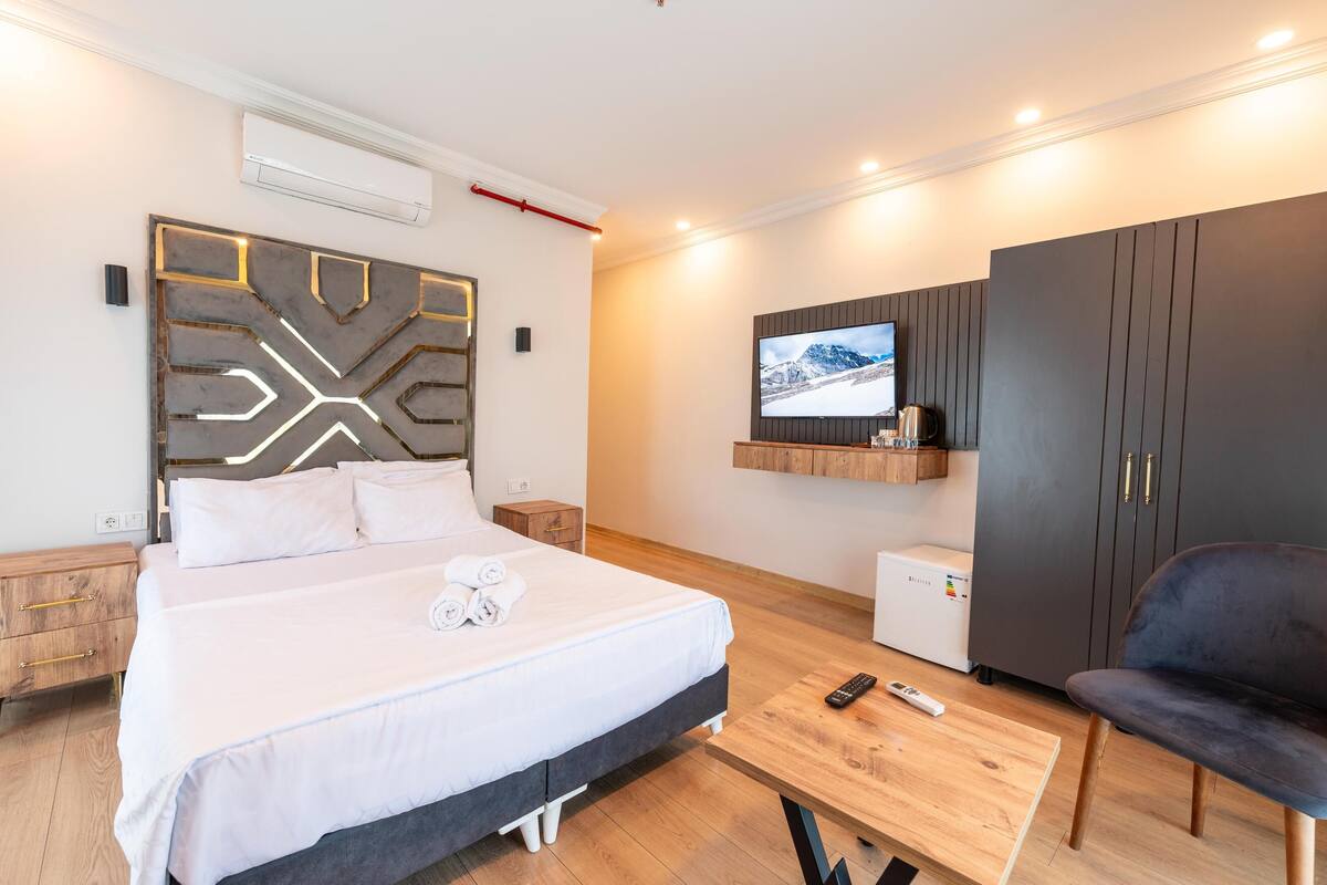 A modern hotel room with a large bed, a TV mounted on the wall, a wardrobe, and a small table with a remote control.