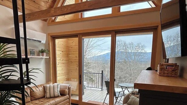 A cozy cabin interior with a wooden ceiling, a sofa, ottomans, a cowhide rug, and a view of a deck and mountains.