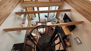 A high-ceilinged room with a wooden interior, a central staircase, and a view of the outdoors through large windows.