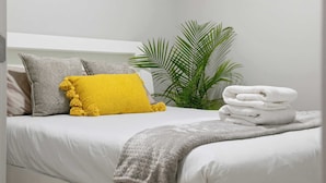 Cama bem arrumada com roupa de cama branca, almofada cinza e amarela, e manta cinza.