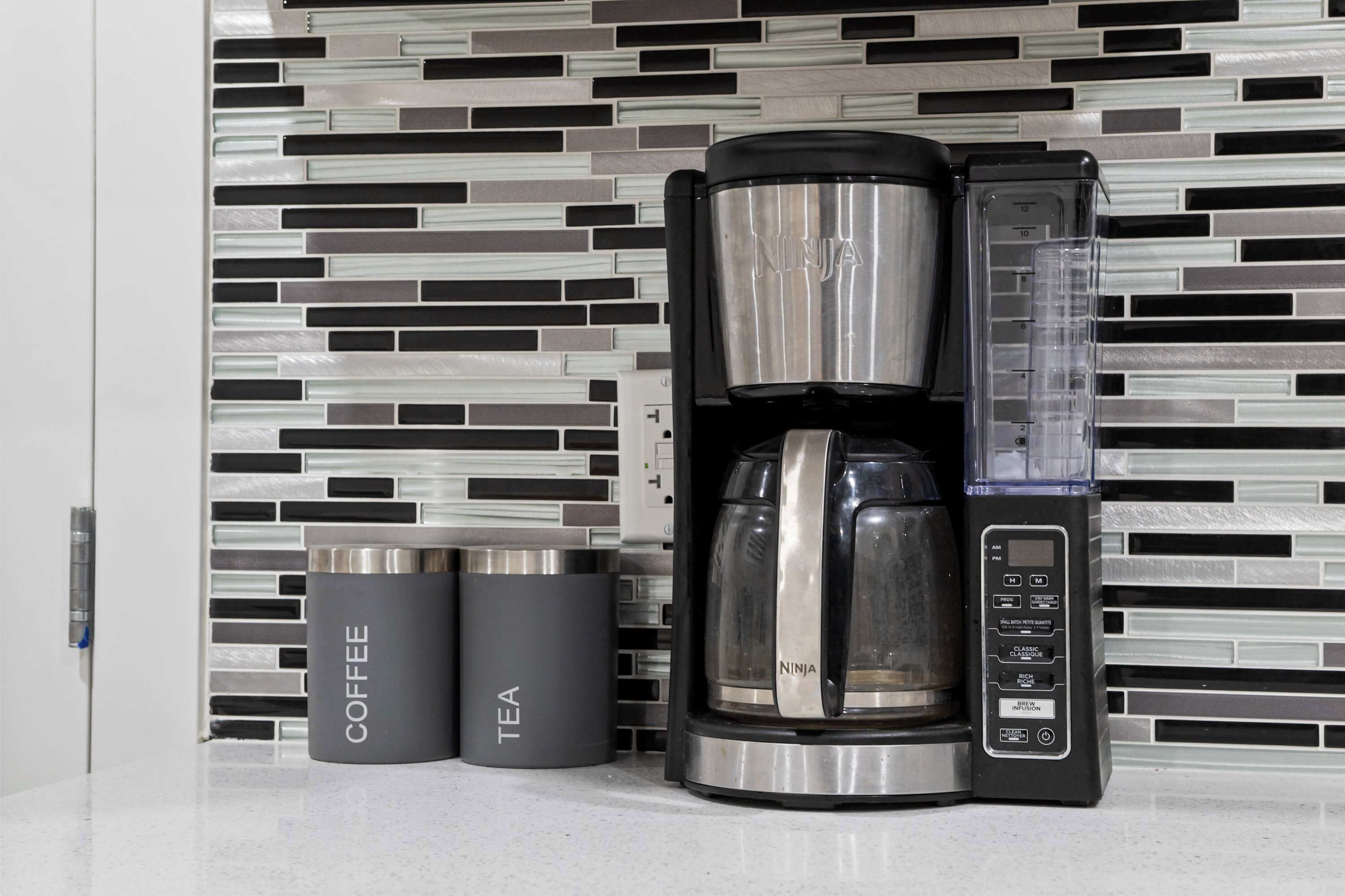 A coffee maker with a digital interface, two labeled containers for coffee and tea, and a modern tiled backsplash.