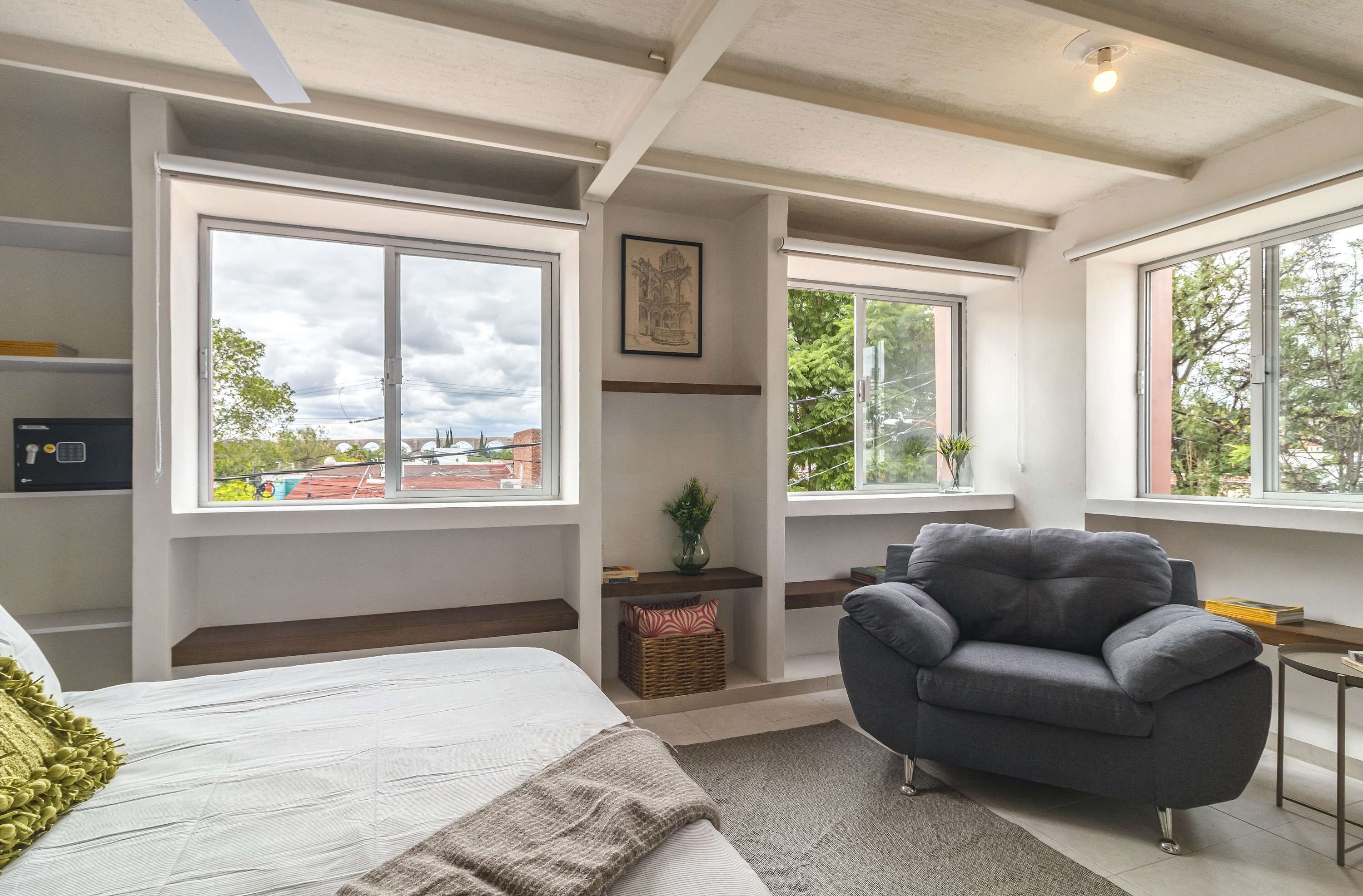 A modern bedroom with a bed, a grey armchair, and large windows offering views of the outside.