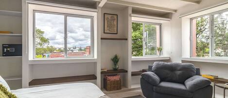 A modern bedroom with a bed, a grey armchair, and large windows offering views of the outside.
