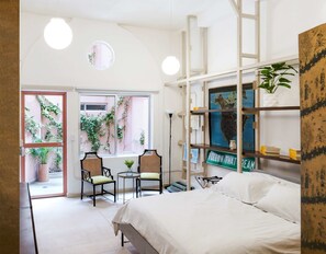 Un dormitorio con una cama, dos sillas, una mesita, una estantería con libros y un mapa, y una ventana con plantas.