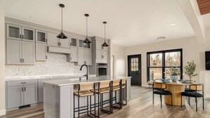 A modern kitchen with grey cabinets, a large island with bar stools, pendant lighting, and a dining area with a round table and chairs. - The Silversands Lakehouse | Hot Tub • Sauna • Game Room (Lansing)