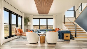 A spacious living room with a large window, a blue sofa, two armchairs, a wooden coffee table, and a staircase with a railing. - The Silversands Lakehouse | Hot Tub • Sauna • Game Room (Lansing)