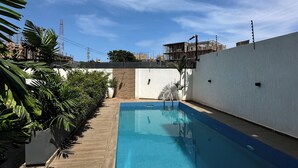 Une piscine rectangulaire entourée d’une terrasse en bois, un mur blanc d’un côté et, au loin, une vue sur un chantier de construction.