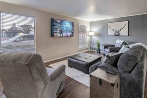 Ein Wohnzimmer mit grauem Sofa, einem schwarzen Hocker, einem an der Wand befestigten Fernseher und einem Bilderrahmen mit dem Abbild eines Stiers.
