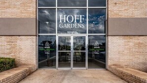 Un edificio con un amplio ventanal de vidrio, el nombre 'HOFF GARDENS' bien visible, y dos logotipos de Airbnb en las ventanas.