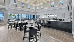 A spacious dining area with multiple tables and chairs, a bar area with stools, and large windows offering views of the outdoors. - Wildblue - William Raveis Rentals (Ft. Myers)