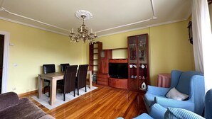Una sala de estar con piso de madera, una mesa de comedor con sillas, un sofá azul, un televisor y una araña.