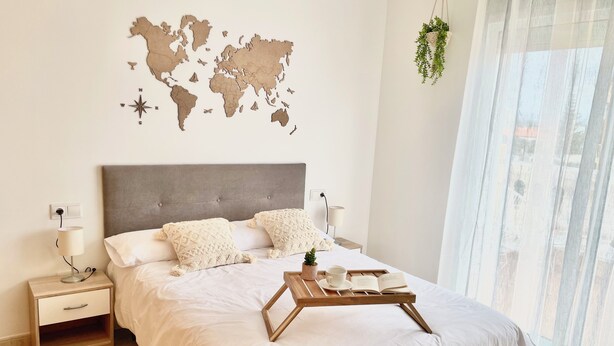 Una cama bien hecha con un mapa mundial en la pared, una mesita de noche con una lámpara y una planta pequeña colgando de la ventana.