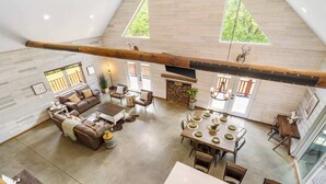 A spacious loft-style living area with a high ceiling, a fireplace, a dining table set for four, and a large window overlooking greenery.
