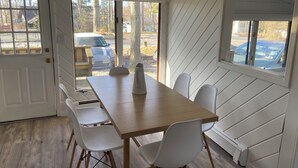 A dining area with a wooden table and white chairs, large windows, and a view of parked cars outside. - Cozy Cottage Steps from the Beach in the North Fork (Calverton)