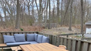 A wooden deck with a long table and chairs, a blue sofa, and a wooden fence. - Cozy Cottage Steps from the Beach in the North Fork (Calverton)