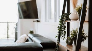 A modern living room with a black sofa, a mounted TV, and a wooden shelf with plants.