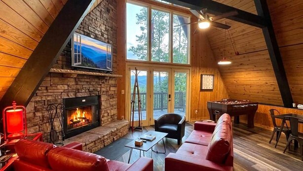 A cozy loft-style room with a stone fireplace, red leather seating, a pool table, and a large window offering a view of the outdoors.