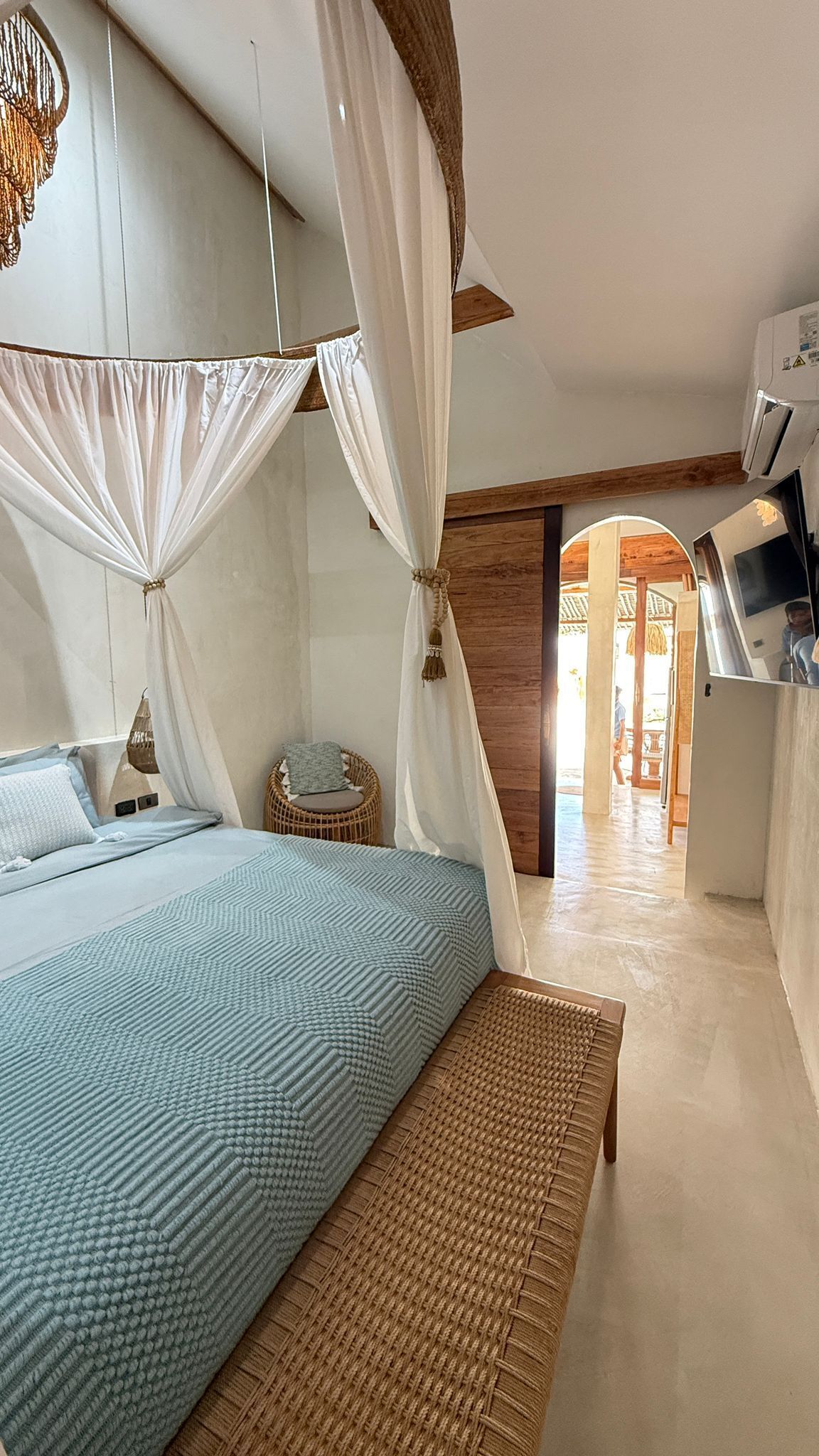 A bedroom with a bed, a bench, a hanging light fixture, and a doorway leading to another room.