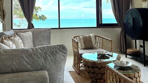 Ein Wohnzimmer mit großem Fenster und Meerblick, einer Couch, einem Korbstuhl, einem Glastisch und einem Lautsprecher.