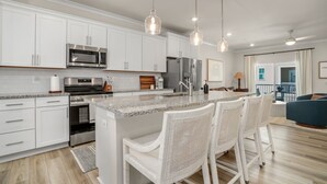 A modern kitchen with white cabinetry, stainless steel appliances, and a granite countertop. There are three white wicker chairs around the island.