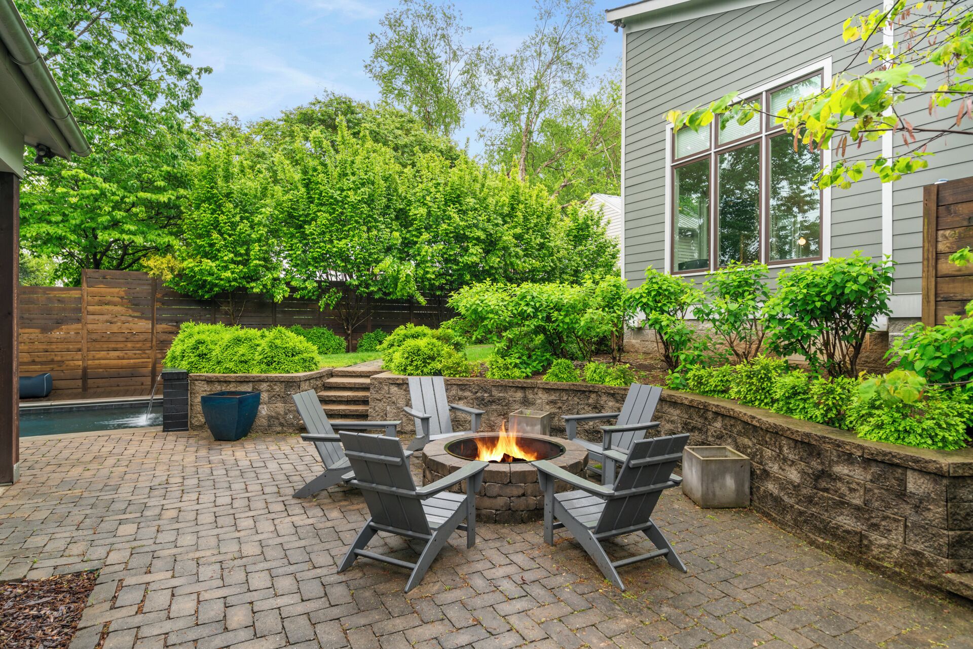 Un patio trasero con un brasero rodeado de sillas Adirondack, un muro de piedra y abundante vegetaci ón.