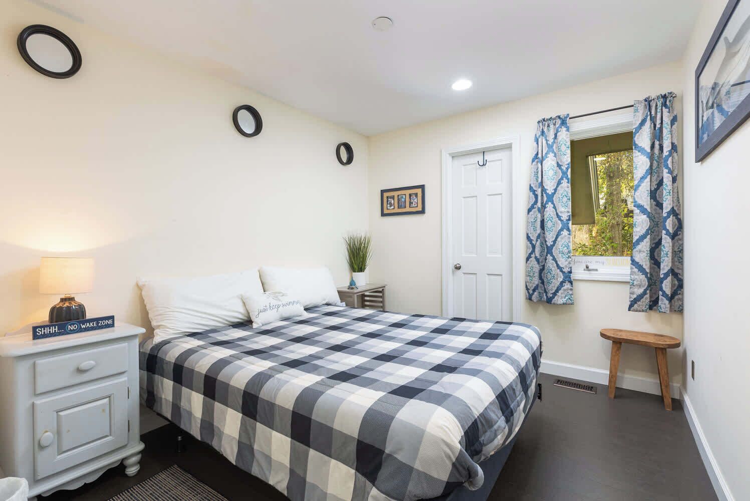 A bedroom with a checkered bedspread, a bedside table with a lamp, a small stool, a door, and a window with patterned curtains.