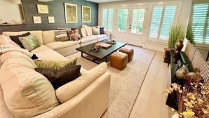 Un salon moderne avec un canapé d’angle, une table basse et une grande fenêtre équipée de stores.