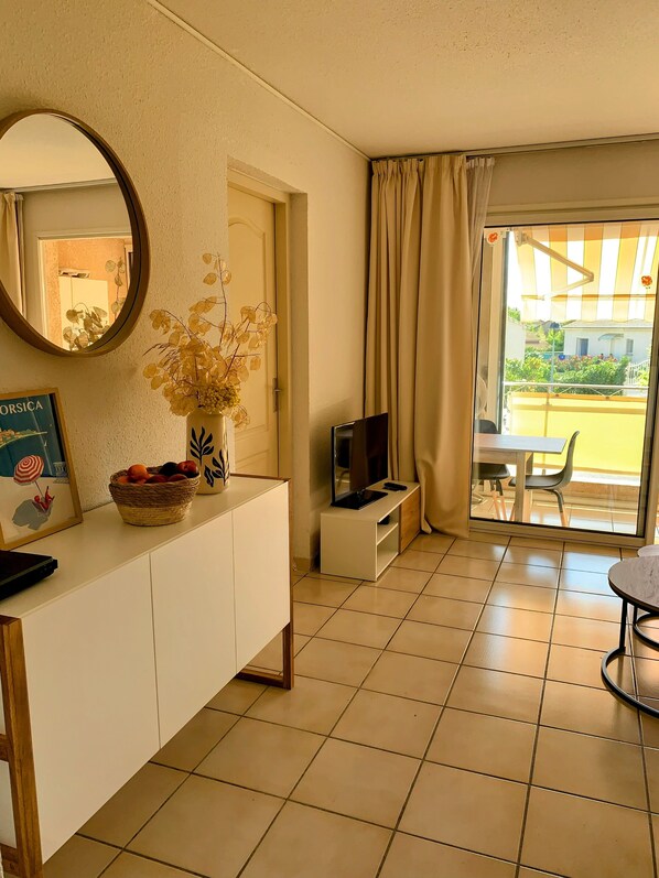 A hotel room with a white cabinet, a mirror, a television, a dining area with a table and chairs, and a view of the outside. - Chez Aline - One-bedroom ground-floor apartment by the sea (Cervione)