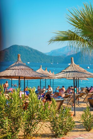 A beach with thatched umbrellas, lounge chairs, and people relaxing. - Hotel Caprea (Marmaris)
