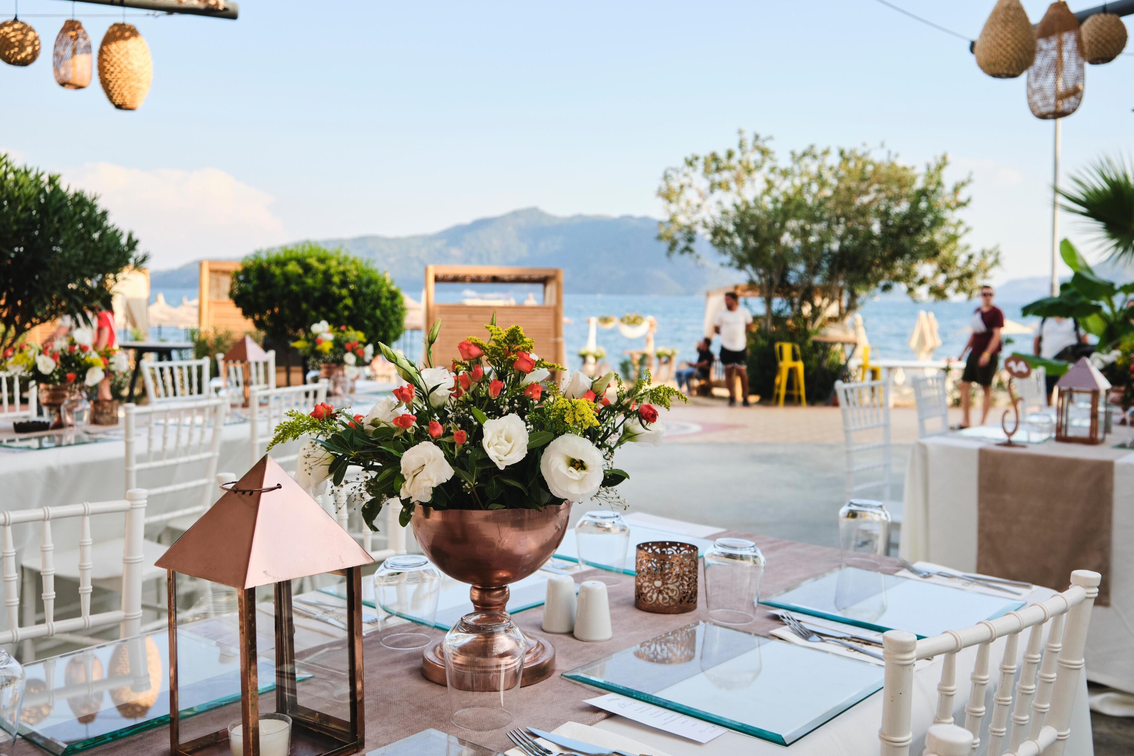 An outdoor dining area with tables set for a meal, a copper bowl with flowers, and a view of the sea and mountains in the background.