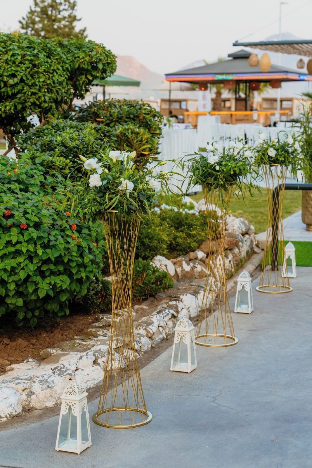 Terraza al aire libre con mesas y sillas blancas, rodeada de follaje y faroles blancos.