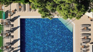 Eine Vogelperspektive zeigt einen rechteckigen Swimmingpool, umgeben von Liegestühlen und Tischen, mit Bäumen im Hintergrund.