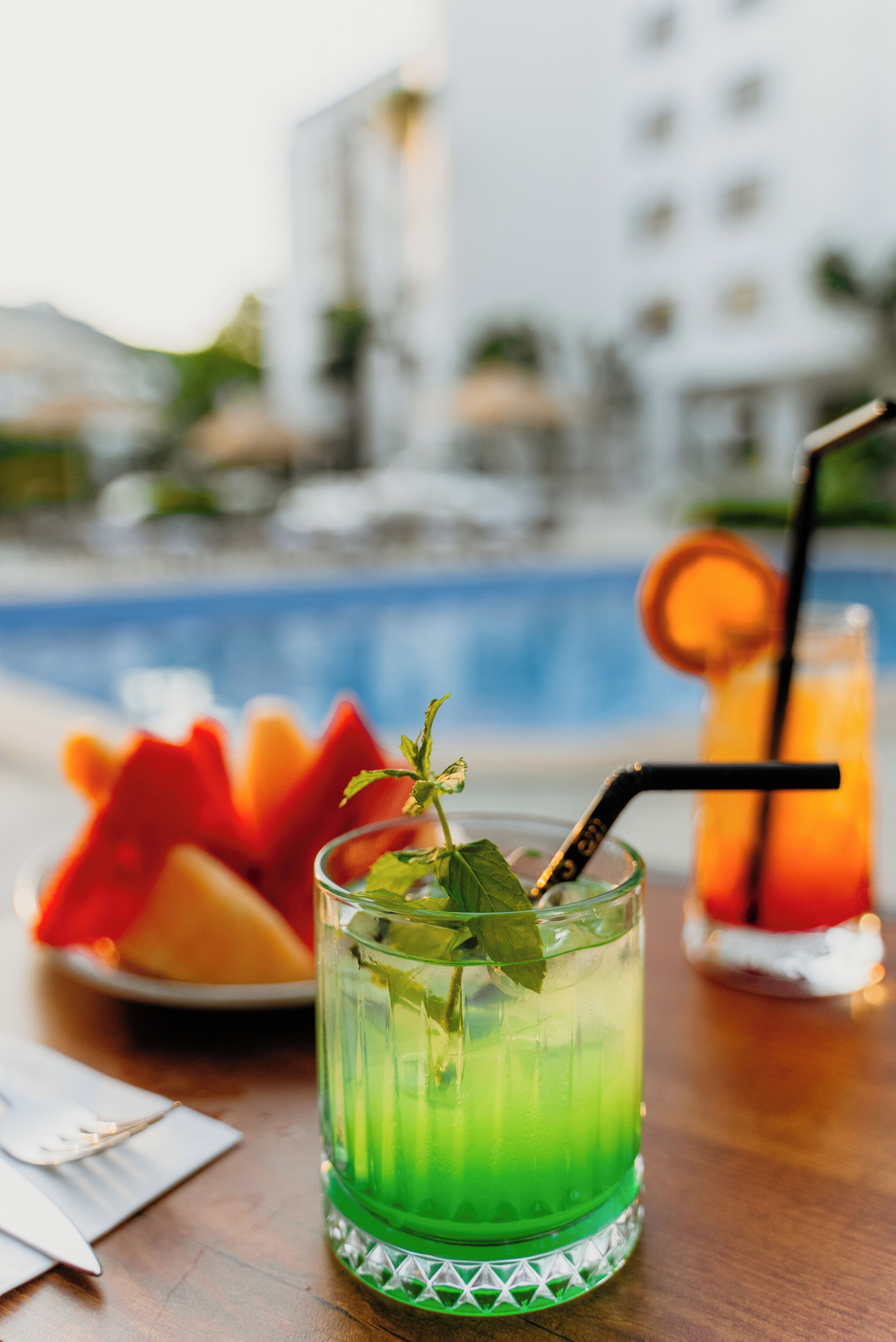 A glass of green cocktail with a mint garnish, a slice of watermelon, and a black straw on a wooden table.