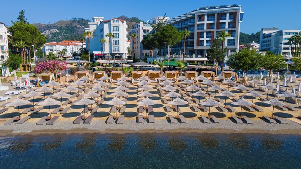 A beachside hotel with numerous sun loungers and umbrellas, a pool, and a view of the surrounding buildings and hills. - Hotel Caprea (Marmaris)