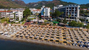 Ein Küstenhotel mit mehreren Gebäuden, einem Strand mit Liegestühlen und Sonnenschirmen sowie einer Bergkulisse.