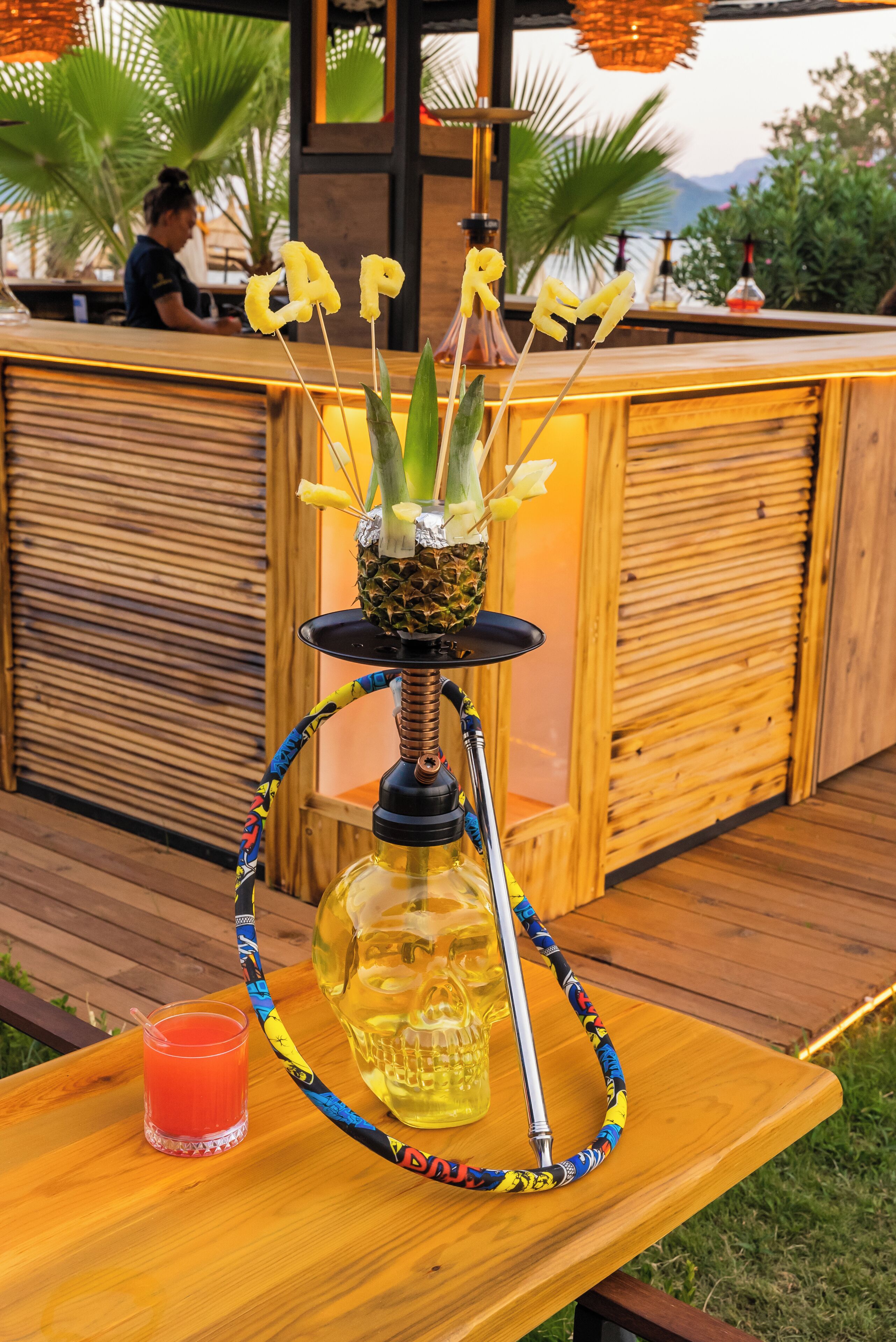 Una barra de madera con un narguilé decorado con piña y flores, un vaso de bebida roja, y una vista de una persona al fondo.