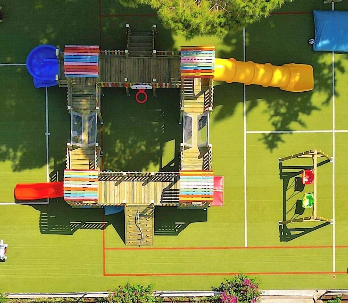 An aerial view of a playground with various play structures, including slides and climbing areas, surrounded by a fenced area.