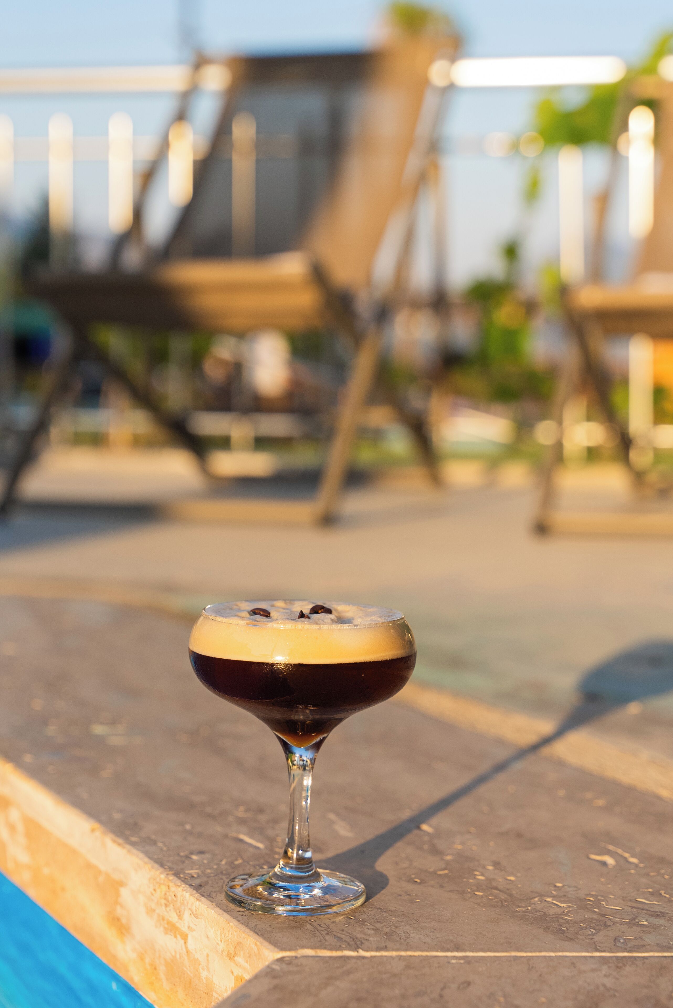 A cocktail glass on a terrace with a pool and lounge chairs in the background.