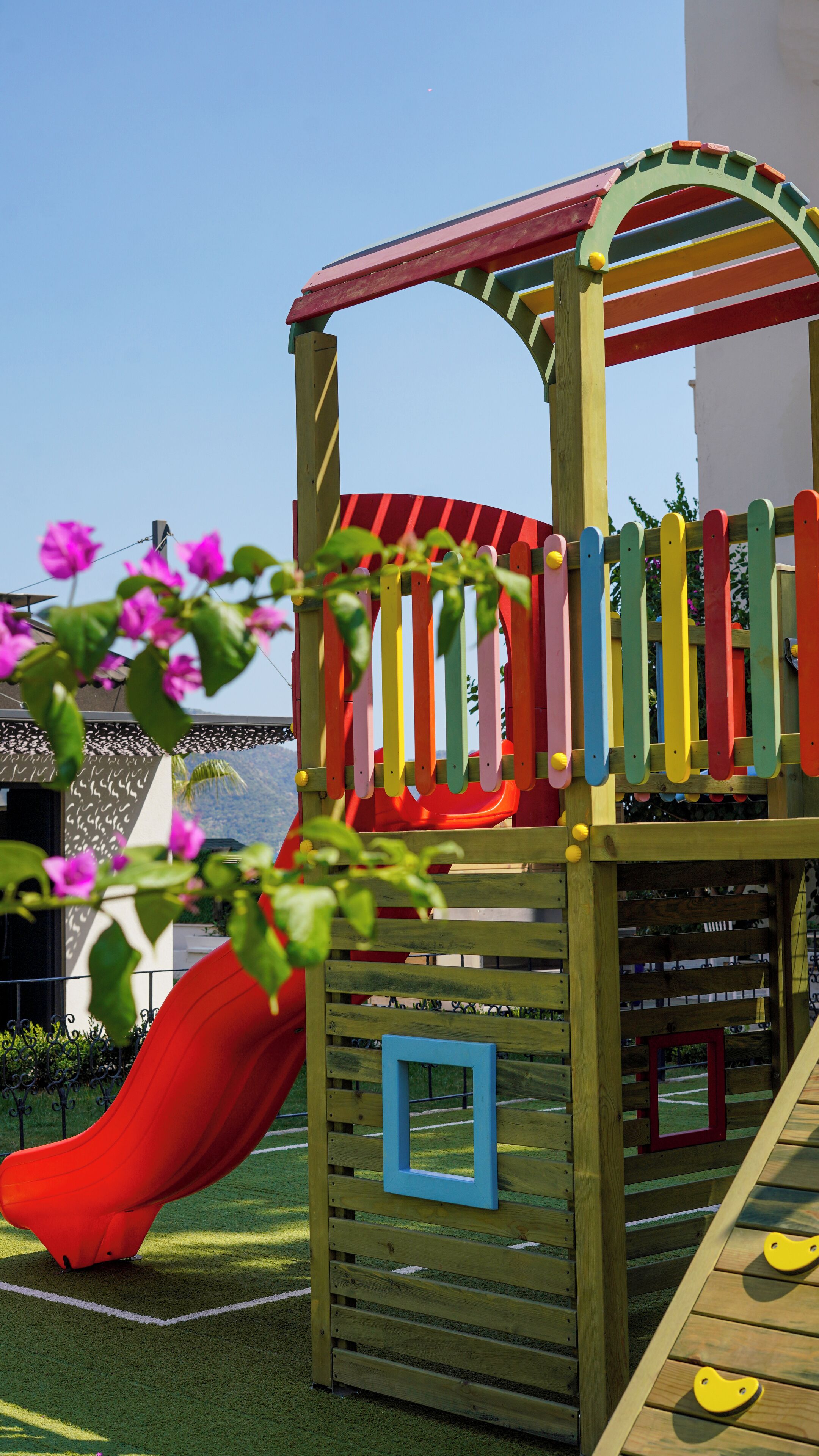 A wooden play structure with a red slide, surrounded by greenery and flowers.