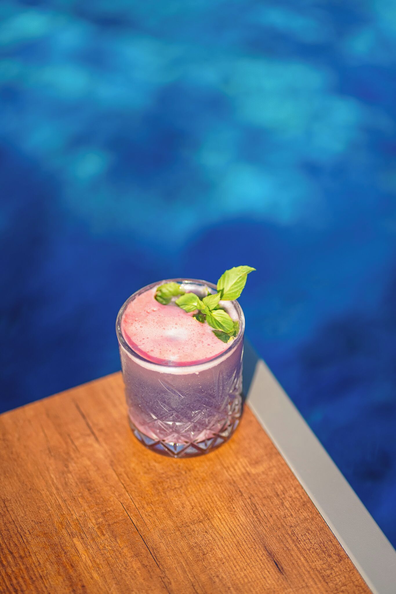 A glass of pink drink with a mint garnish on a wooden table by a pool.