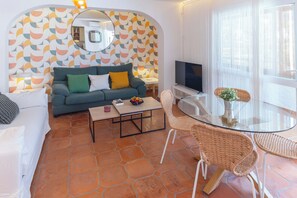 A living room with a sofa, coffee table, and wicker chairs. A TV is mounted on the wall, and a window with sheer curtains is visible. - ¡A pie de playa! Apartamento Cap Negret (Altea)