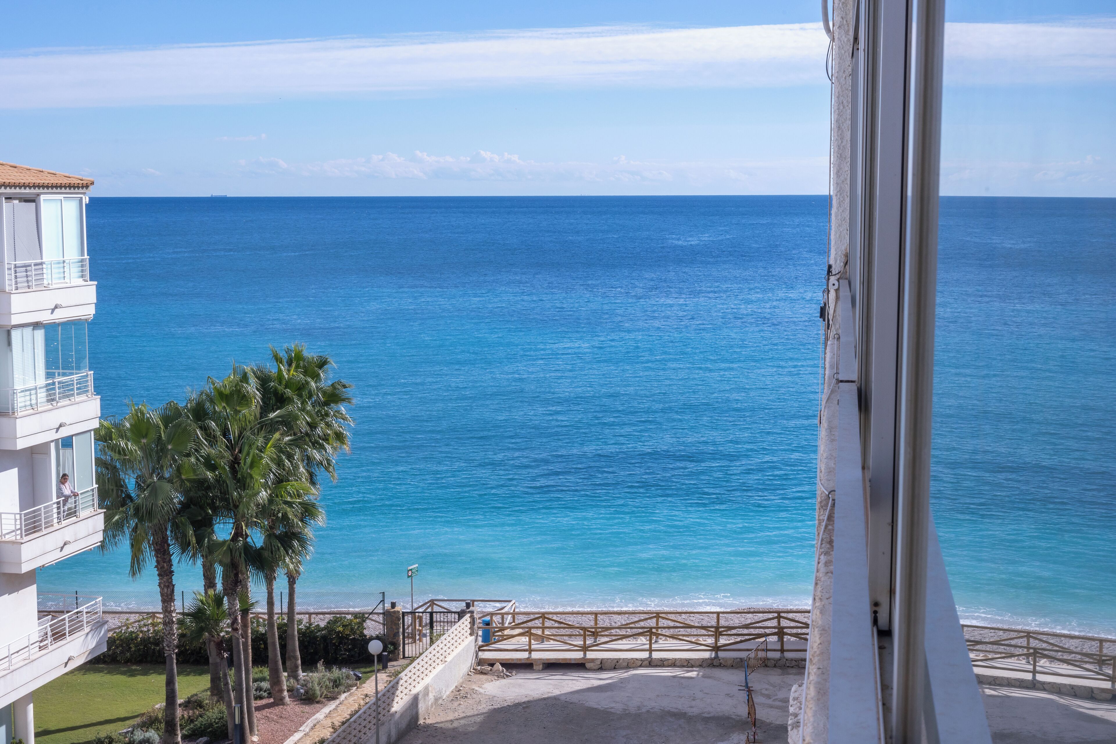 A coastal view from a balcony overlooking the sea, with a building and palm trees in the foreground.