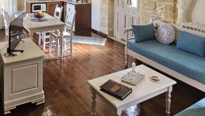 A spacious living area with a blue sofa, a white coffee table, and a dining table with chairs. There is a kitchen in the background with wooden cabinets and a stone wall. - Arhontiko Arhanes - Cretan Fairytale Gem With Pool (Archanes-Asterousia)