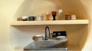 Une cuisine avec un évier en pierre, une étagère en bois où sont disposées diverses tasses et verres, et un mur incurvé.