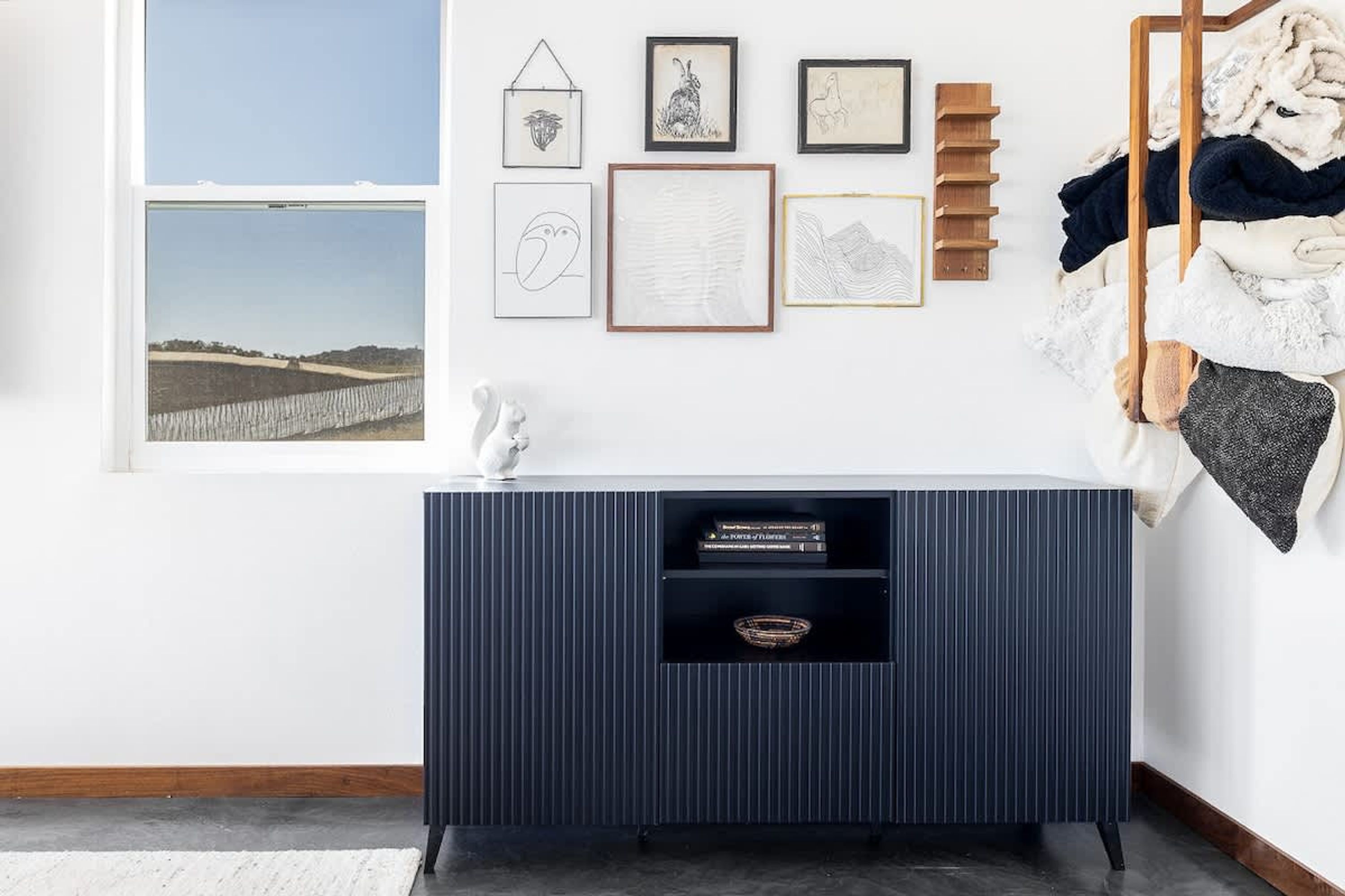Une pièce dotée d’une grande fenêtre, d’un buffet à rayures bleu foncé et de divers tableaux encadrés accrochés au mur.