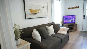 Una sala de estar con un sofá gris oscuro, una mesita de madera con una planta, un televisor sobre un mueble de madera y un cuadro de un barco en la pared.