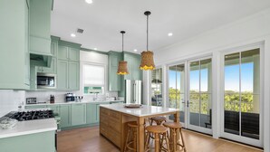 Una cocina moderna con armarios verde claro, una isla central con taburetes y un amplio ventanal corredero que da a un balcón.