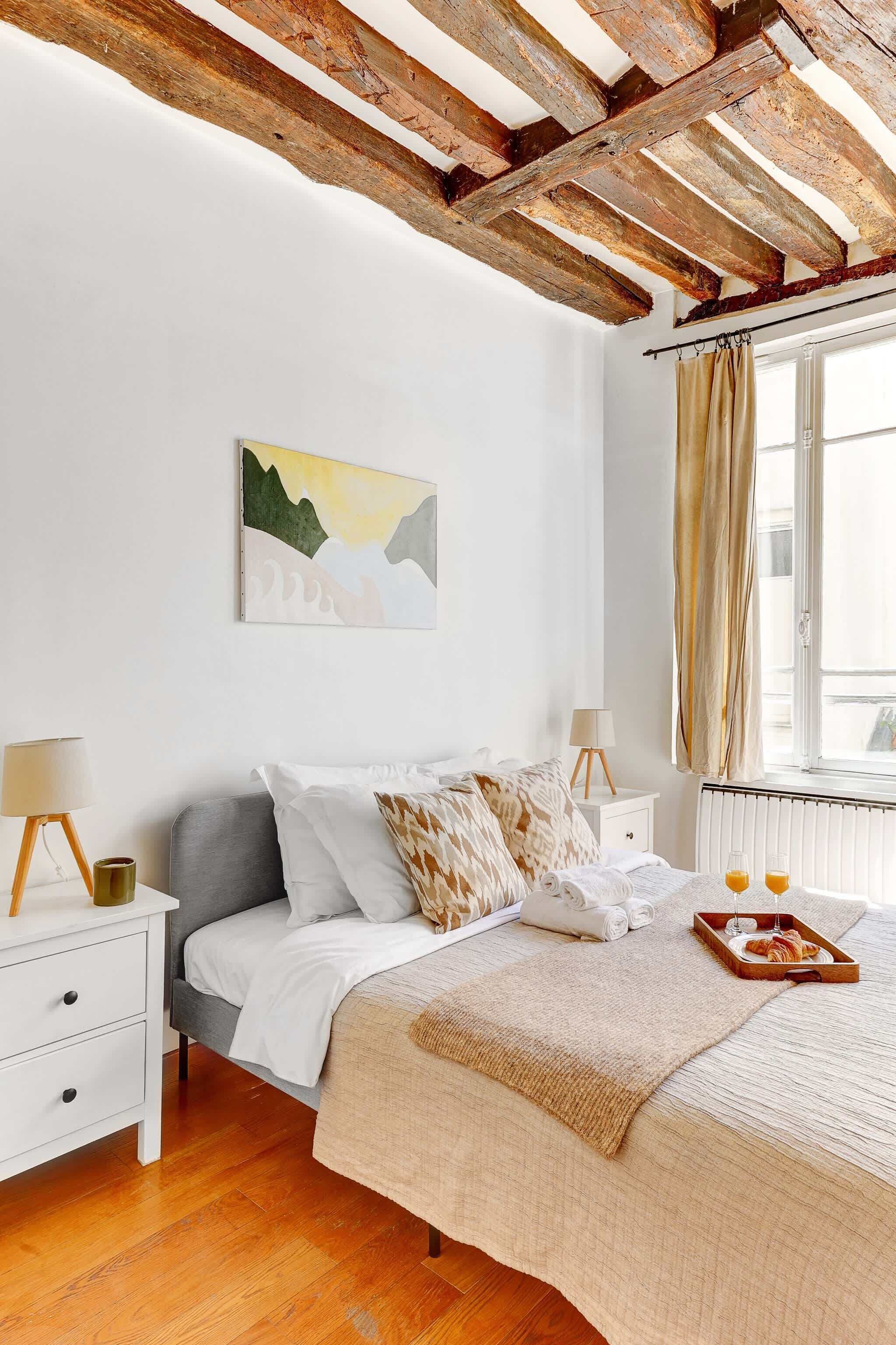 Ein Schlafzimmer mit einem Bett, Nachttischen, einem Heizkörper, einem Fenster mit Vorhängen und einem Bild an der Wand.
